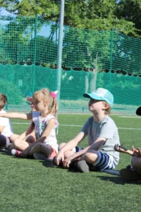 grupa dzieci siedzących na boisku szkolnym podczas letnich zajęć sportowych
