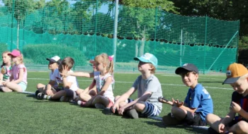 grupa dzieci siedzących na boisku szkolnym podczas letnich zajęć sportowych